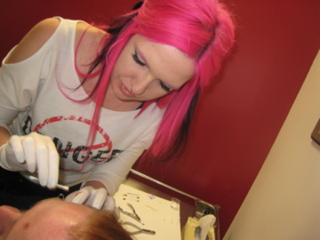 A teenage girl with pink hair and white gloves holds a q-tip to a persons face. She appears to be cleaning an eyebrow ring on their face.